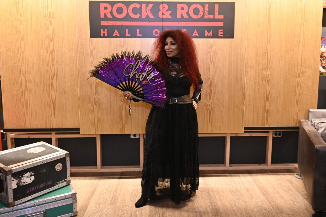 Chaka Khan stands in front of the Rock and Roll Hall of Fame sign with a purple fan wearing a black dress