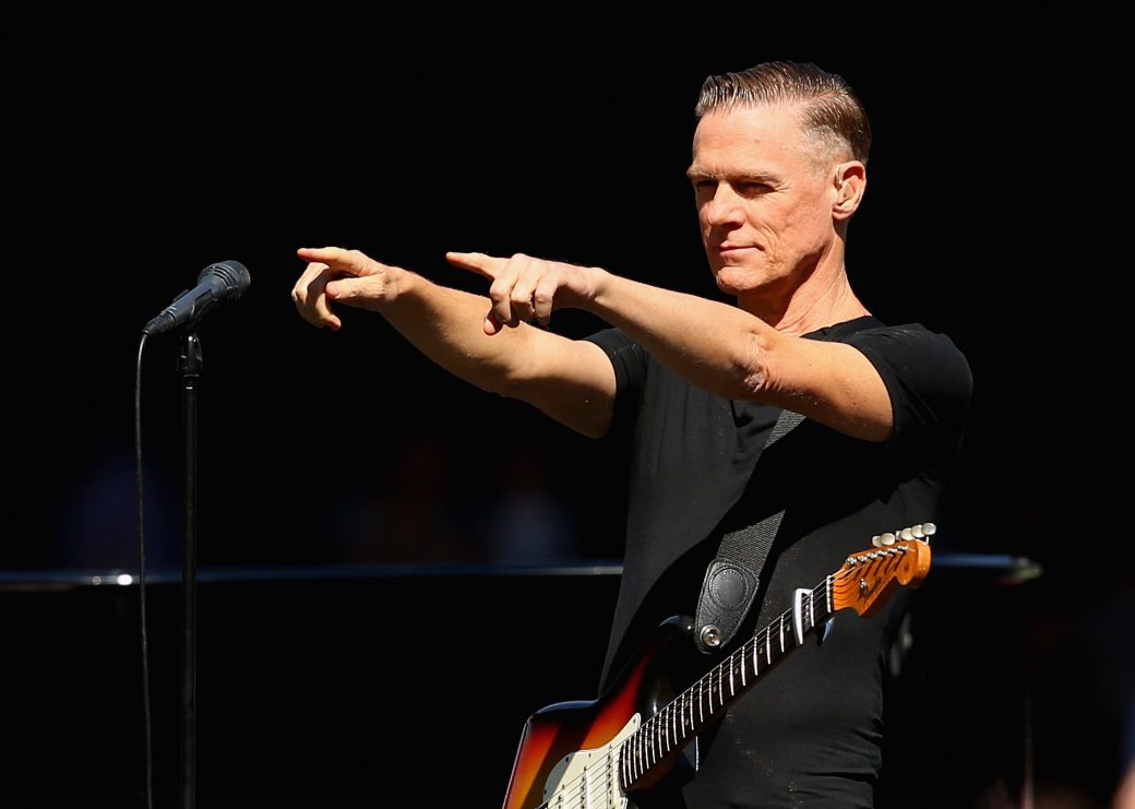 Bryan adams pointing fingers at the audience behind a microphone wearing a black tee with his guitar strapped on