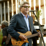 steve miller wearing sunglasses playing guitar in front of a mic